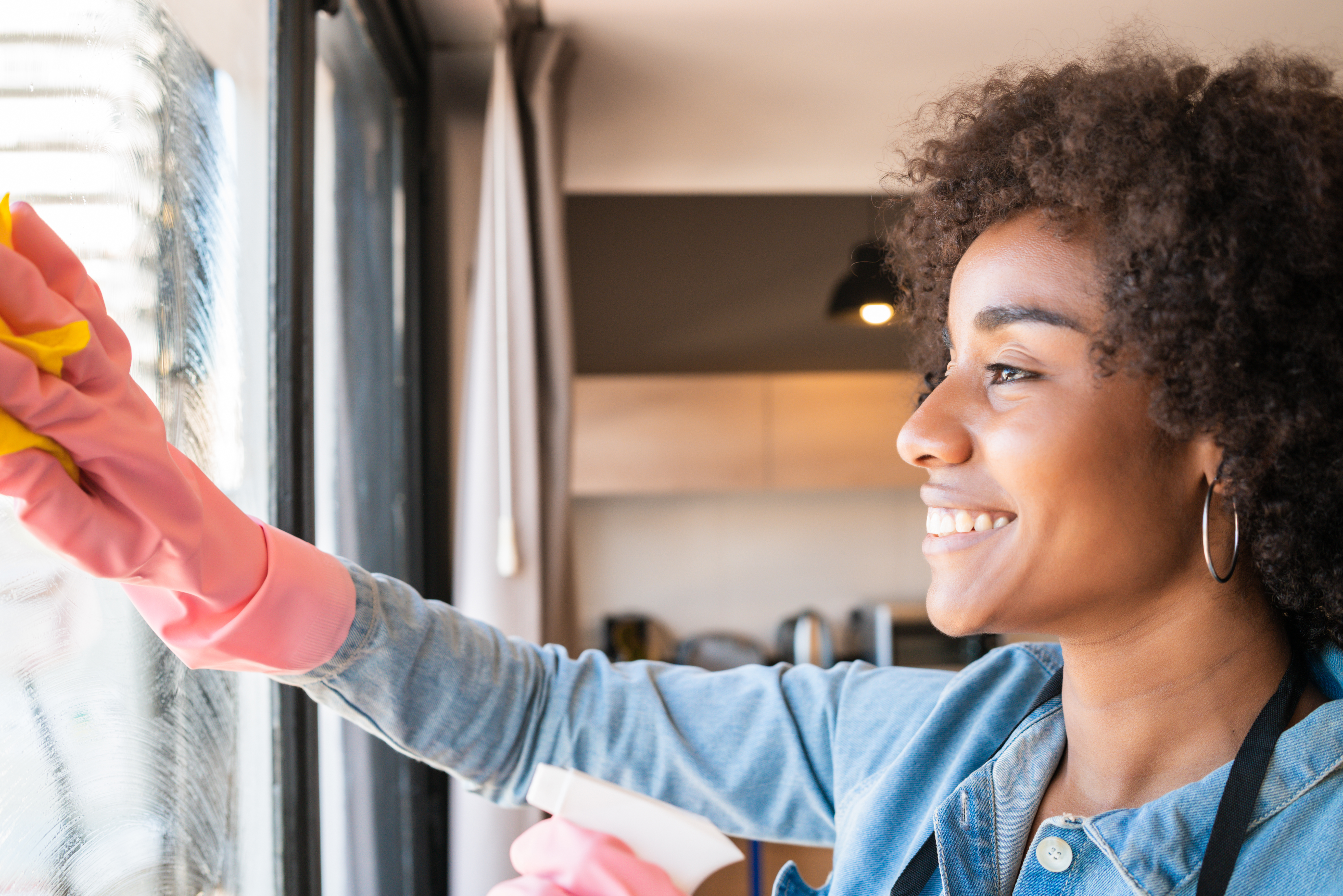 Portrait of young afro woman in gloves cleaning window with rag at home. Housework, housekeeping and cleaning concept.