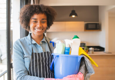 Portrait of young afro woman holding a bucket with cleaning items at home. Housekeeping and cleaning concept.