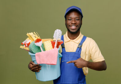 smiling holding and points at bucket of cleaning tools young africanamerican cleaner male in uniform with gloves isolated on green background