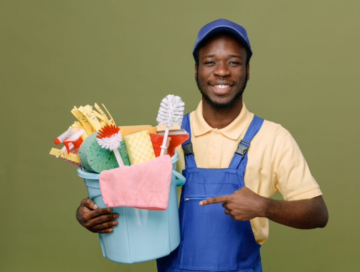 smiling holding and points at bucket of cleaning tools young africanamerican cleaner male in uniform with gloves isolated on green background