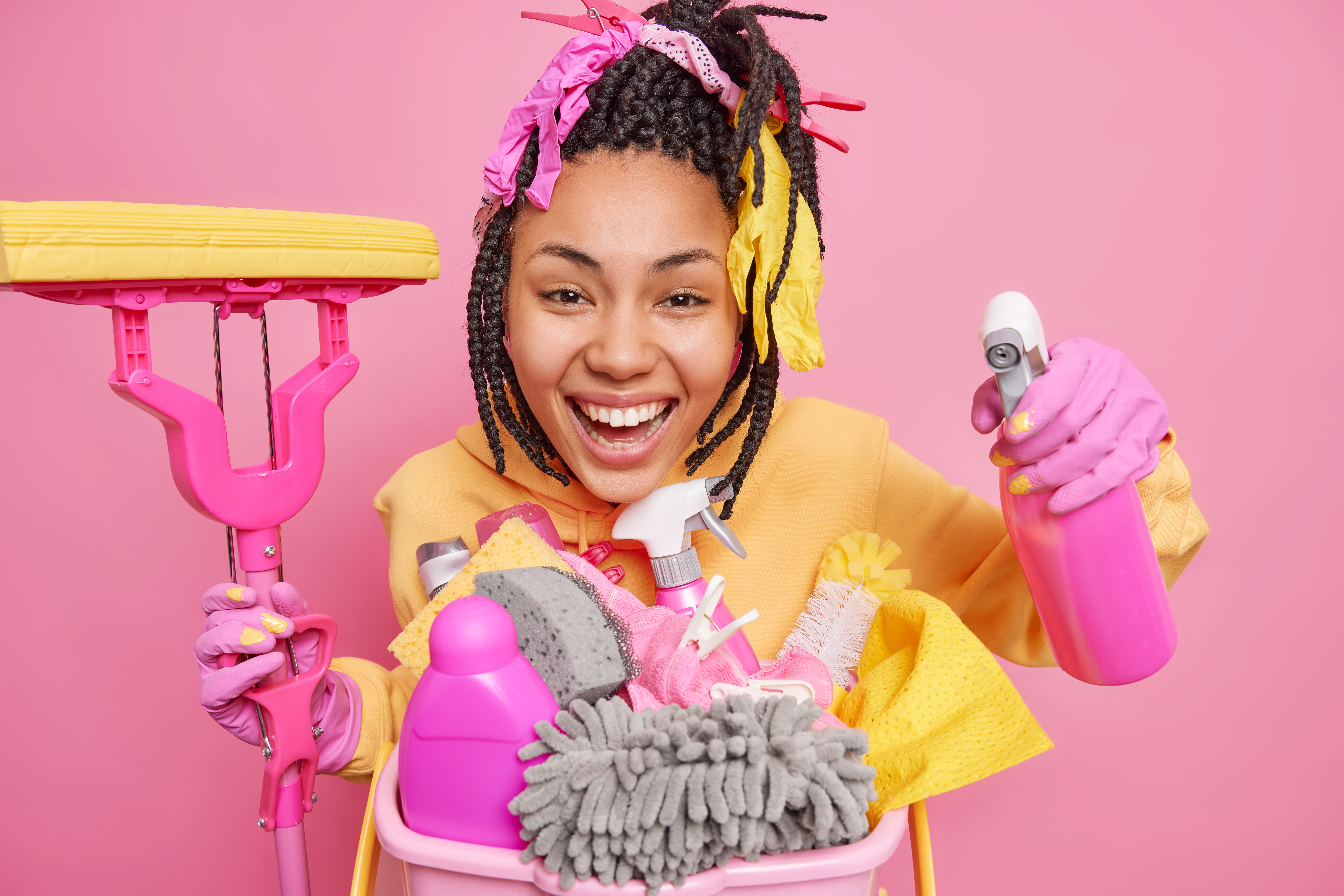 Studio shot of positive dark skinned woman cleans apartment smiles gladfully holds mop and cleaning detergent satisfied with results isolated over pink background. Housework and chores concept